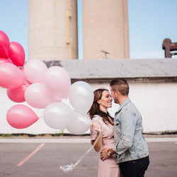 Man hugging Expecting Wife holding Pink Ombre Mix Balloon Bouquet with a midwest background of grain buildings